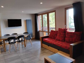A modern living room with a sofa, a dining table with chairs, a flat-screen TV mounted on the wall, and large windows.