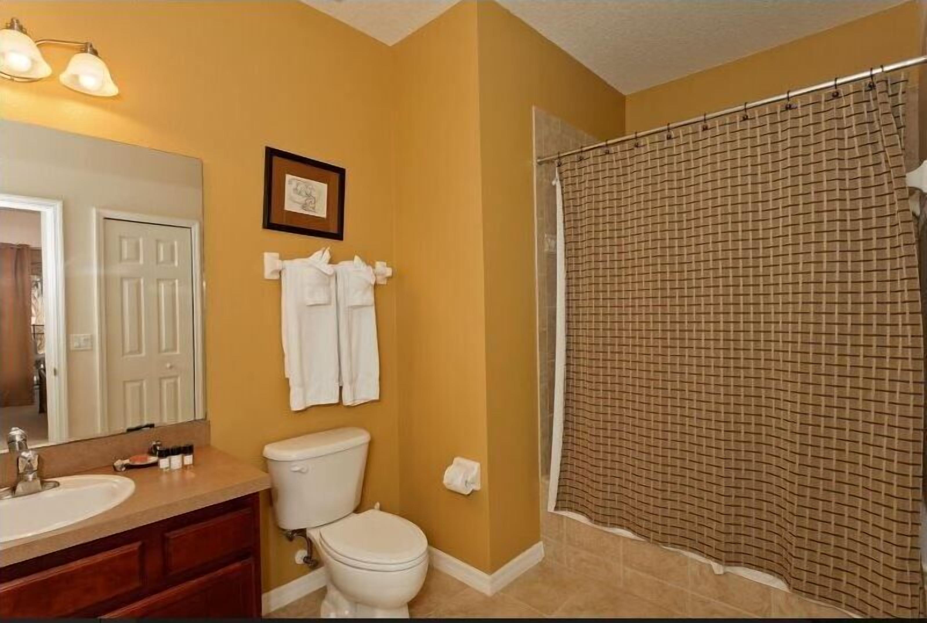 Une salle de bain avec un mur de couleur beige, des toilettes blanches, un lavabo avec un miroir et une douche avec un rideau à carreaux.