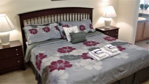 A neatly made bed with floral bedding, two bedside tables with lamps, and a view of a bathroom with a sink and mirror. - Ly218454 - Oak Island Cove - 4 Bed 3 Baths Villa (Kissimmee)