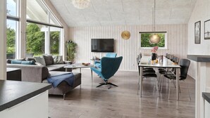 Una sala moderna con un sofá, un sillón azul, una mesa de comedor con sillas, un televisor y una ventana con vistas a zonas verdes.