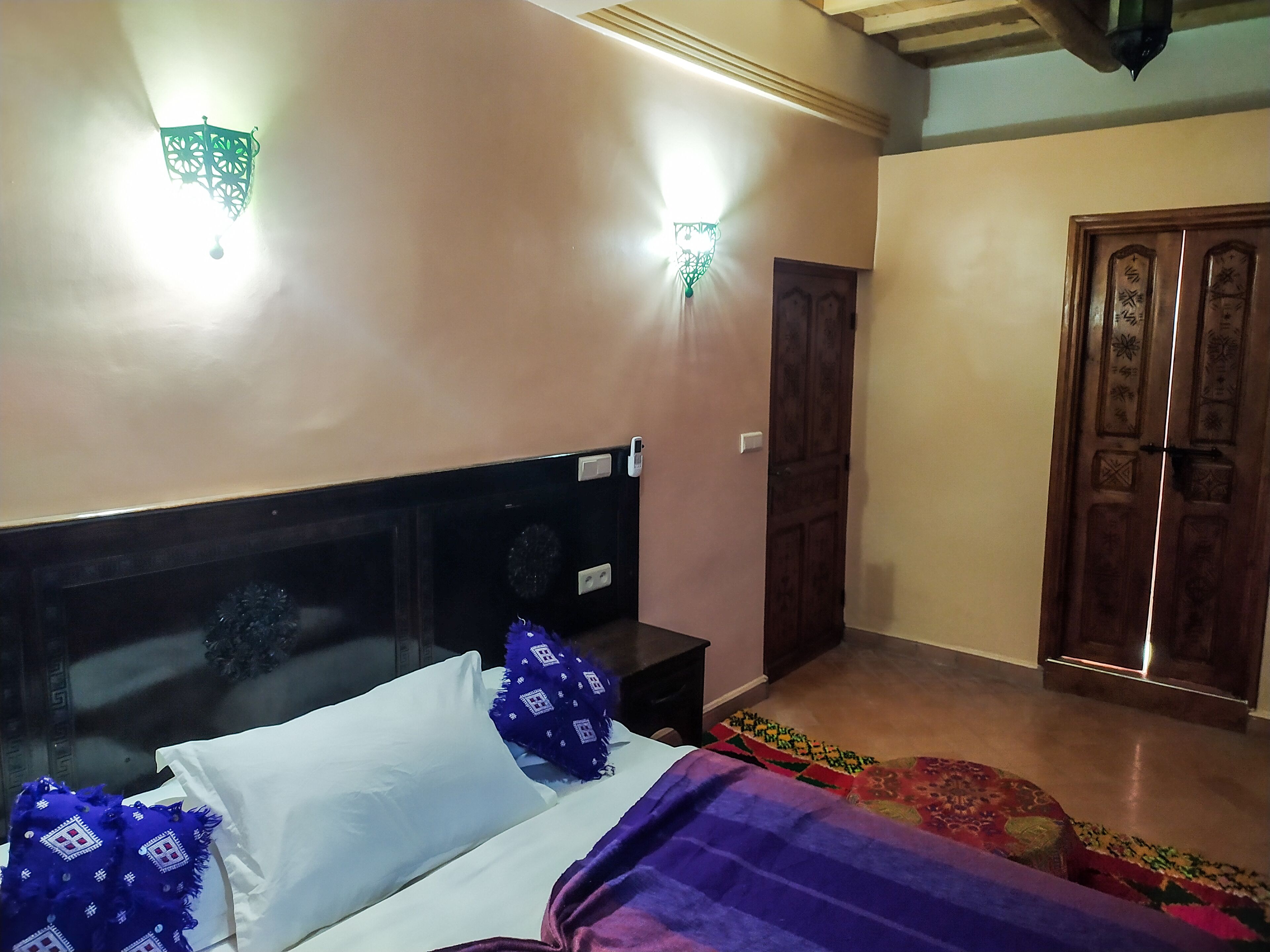 A bedroom with a bed, a nightstand, a wall-mounted light, a door, and a patterned rug.