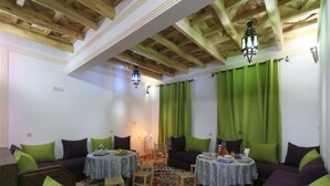 A cozy room with wooden beams, green curtains, and a colorful rug. There are two round tables with chairs, a sofa, and a small table with a candle. - Dar Atlas Charme (Asni)