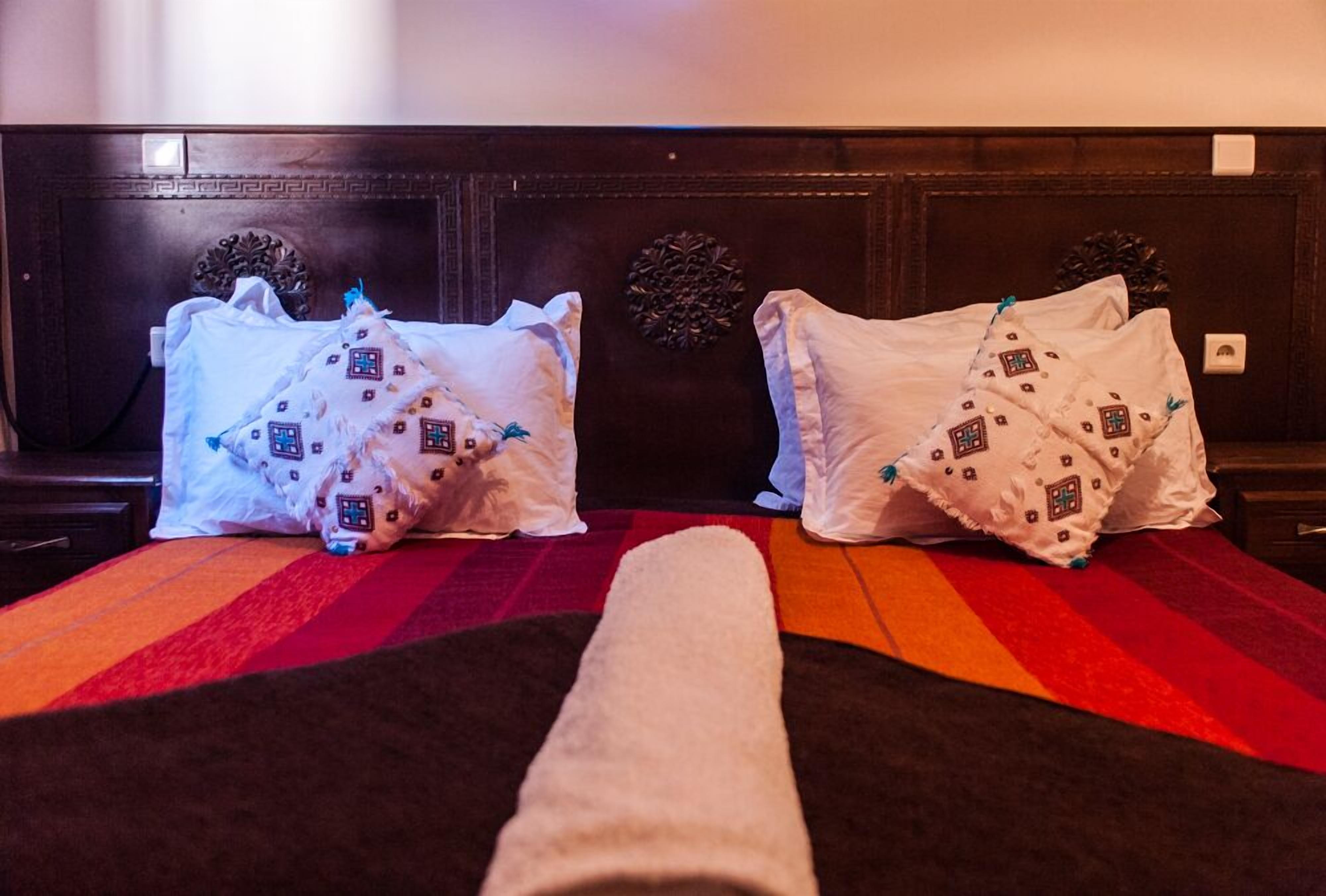 Una camera d'albergo con un letto coperto da una coperta a righe rosse e nere, due cuscini bianchi con motivi decorativi e una testiera in legno finemente intagliata.