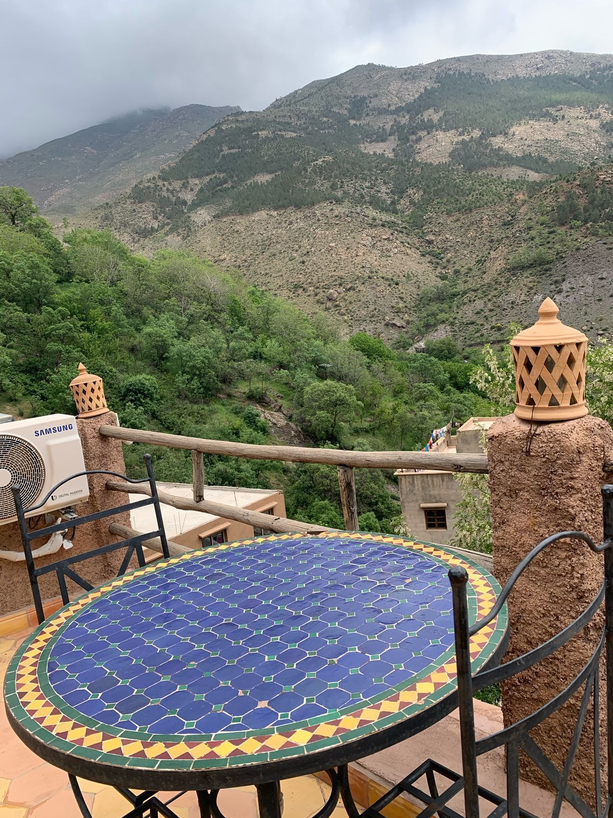 A round table with a blue and yellow patterned tablecloth, a Samsung air conditioner, and a view of a mountainous landscape.