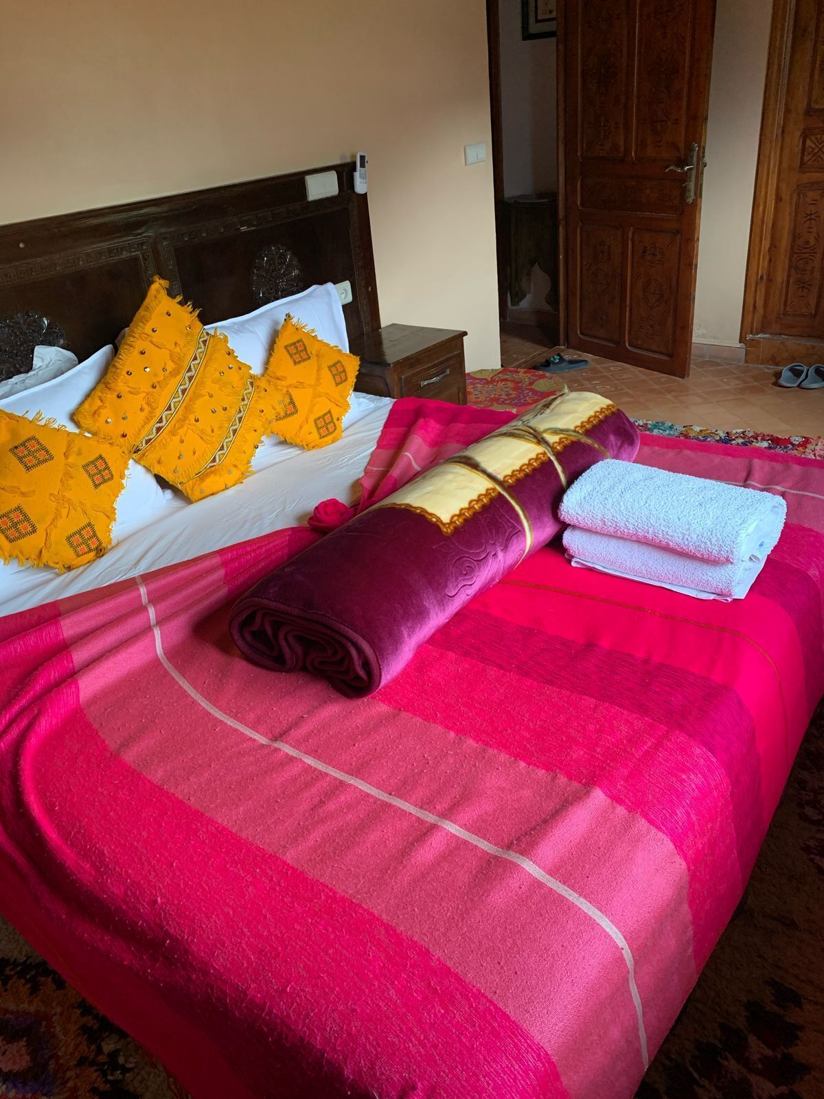 Una cama bien tendida con una colcha a rayas rosada y roja, una manta morada enrollada y dos almohadas amarillas con un estampado de diamantes.