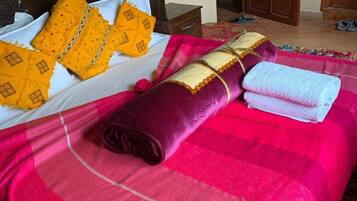 Una cama bien tendida con una colcha a rayas rosada y roja, una manta morada enrollada y dos almohadas amarillas con un estampado de diamantes.