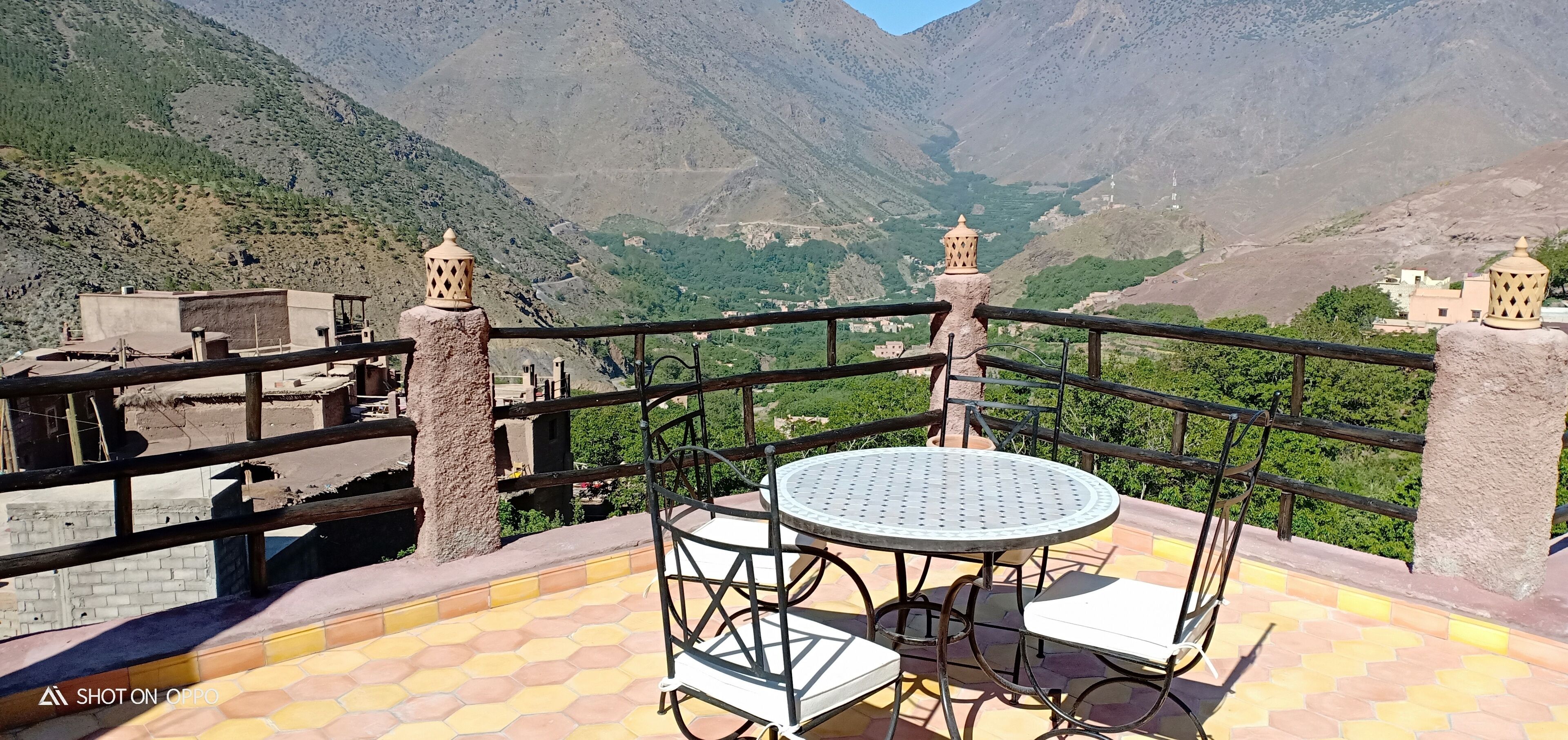 A terrace with a round table and chairs, a railing, and a view of a mountainous landscape.