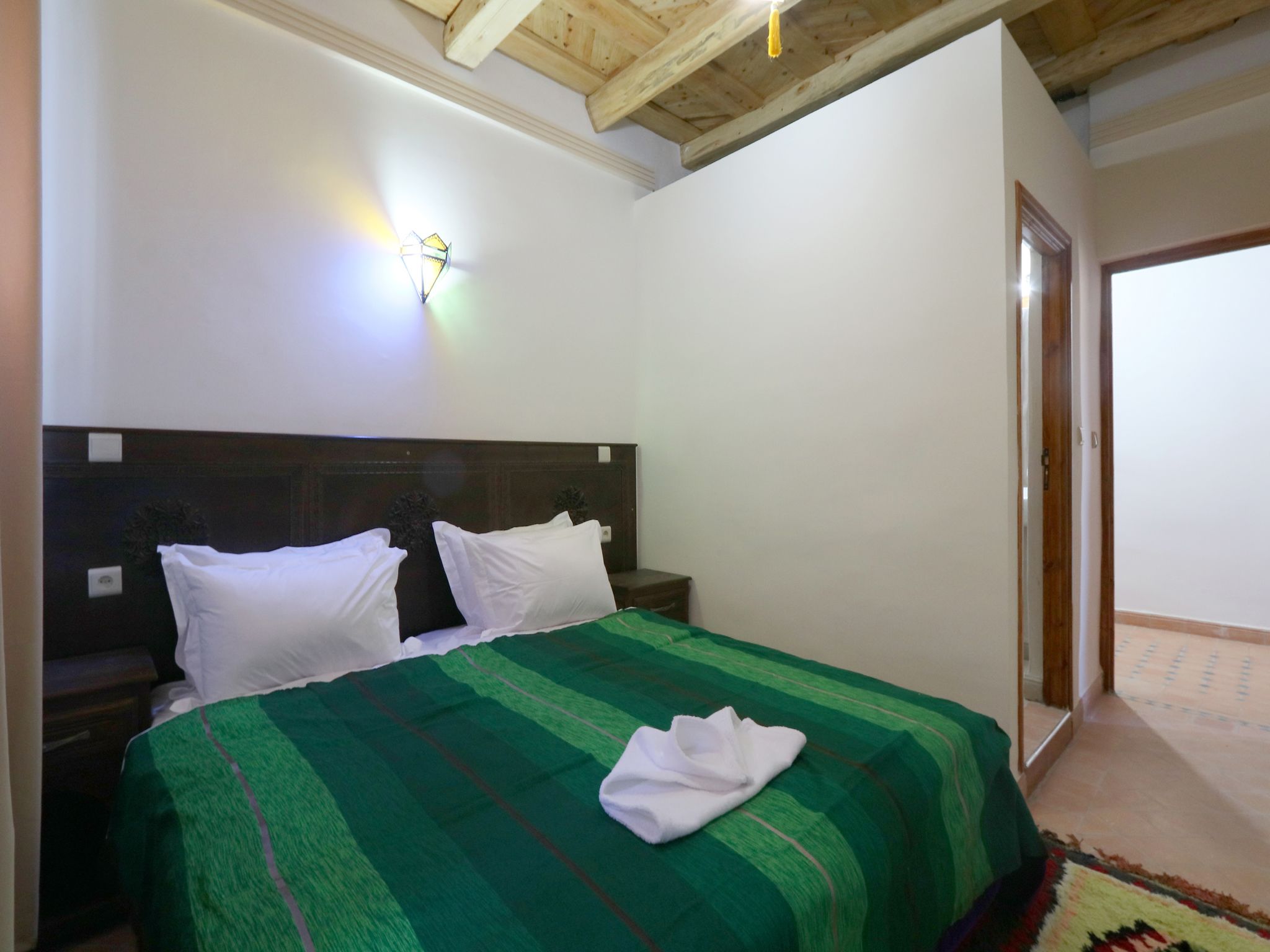Un dormitorio con techo de madera, una cama con colcha verde a rayas, almohadas blancas y una lámpara fijada a la pared.