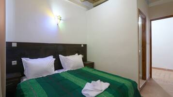 Un dormitorio con techo de madera, una cama con colcha verde a rayas, almohadas blancas y una lámpara fijada a la pared.
