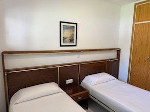 A bedroom with two single beds, a wooden headboard, a bedside table with a drawer, a framed picture on the wall, and a wooden wardrobe. - Agi Rescator 116 Apartamento for rent in Roses Girona. (Roses)