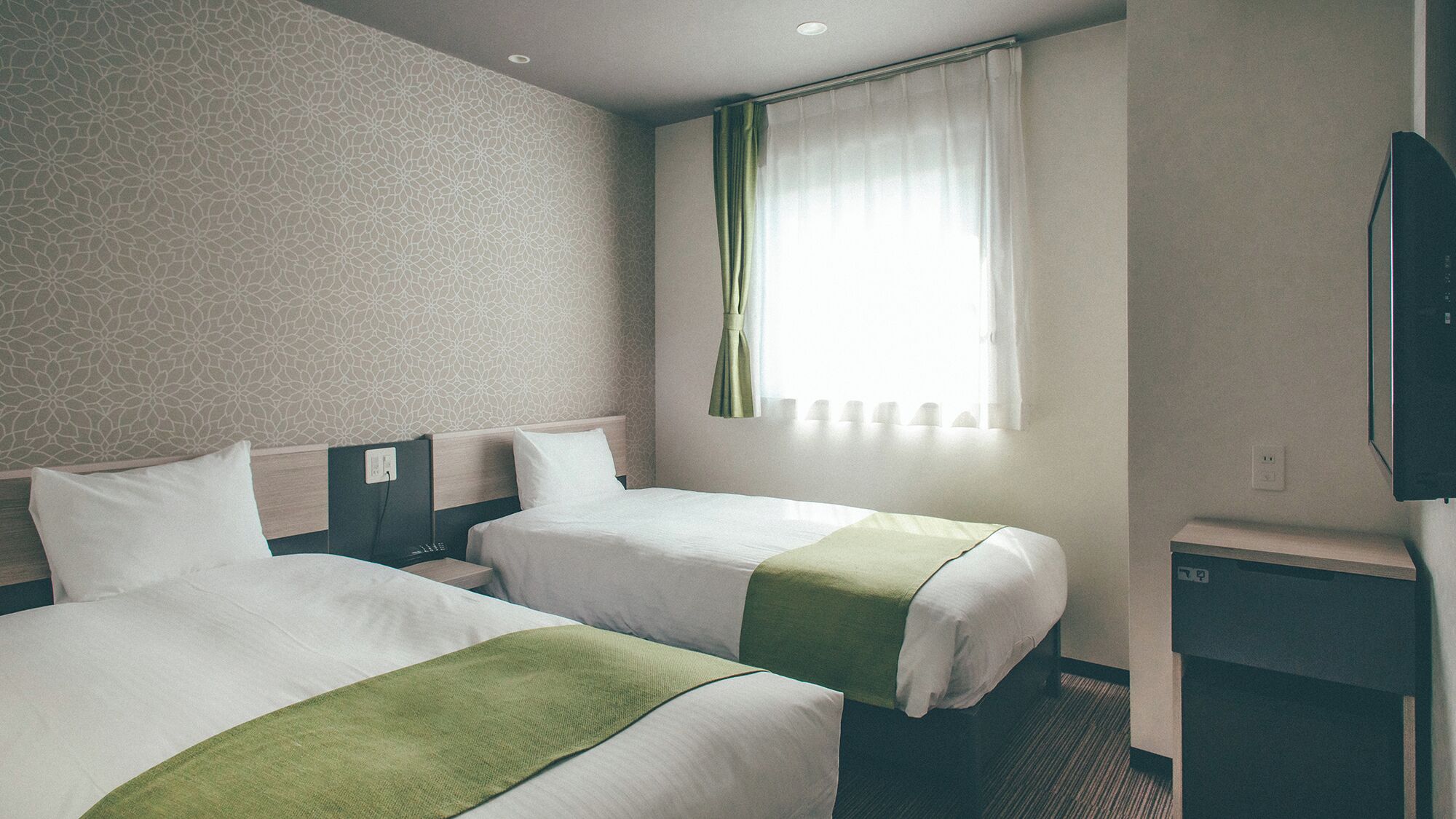 A hotel room with two beds, a television, a window with curtains, and a patterned wall.