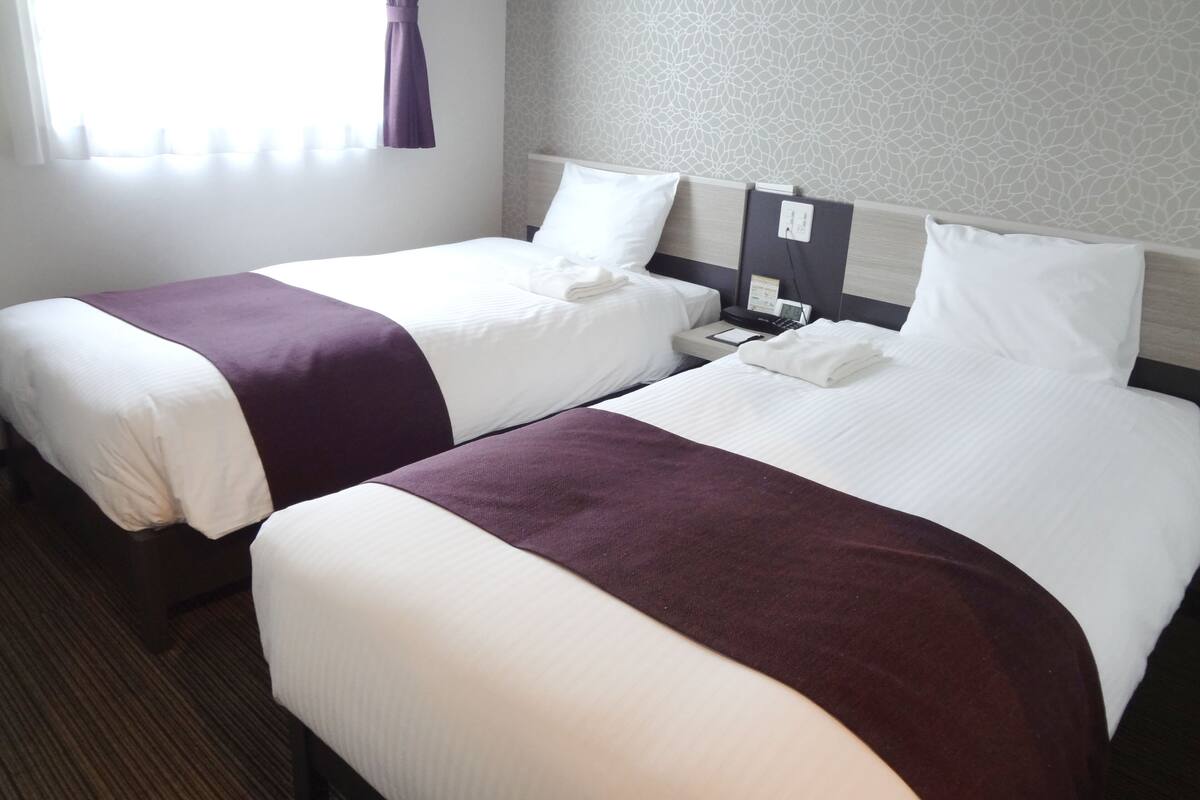 Une chambre d’hôtel avec deux lits simples, chacun recouvert d’une couvre-lit blanche et violette, une table de chevet avec un téléphone et une lampe, et un papier peint à motifs sur le mur.