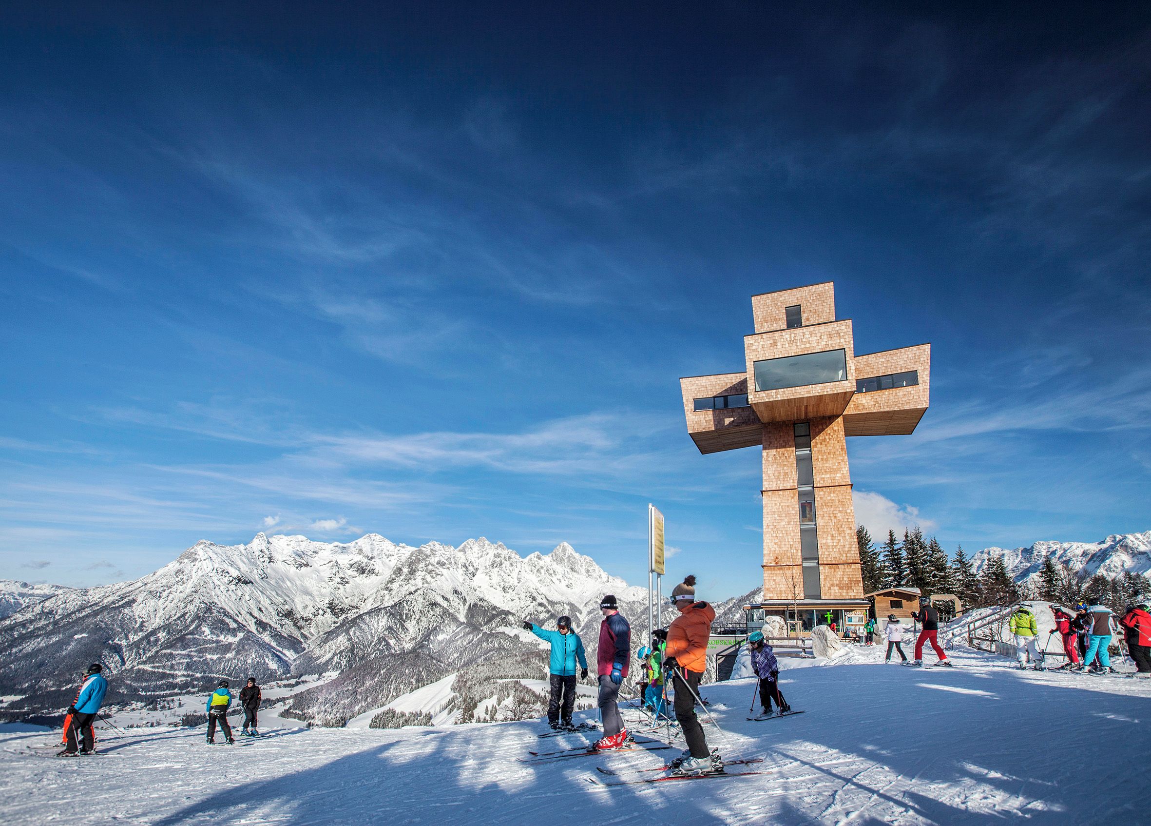 A ski resort with a prominent wooden cross structure and snow-covered mountains in the background.