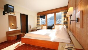 Habitación de hotel con una cama de madera, un televisor montado en la pared, un escritorio con teléfono y una ventana con vistas a un paisaje nevado.