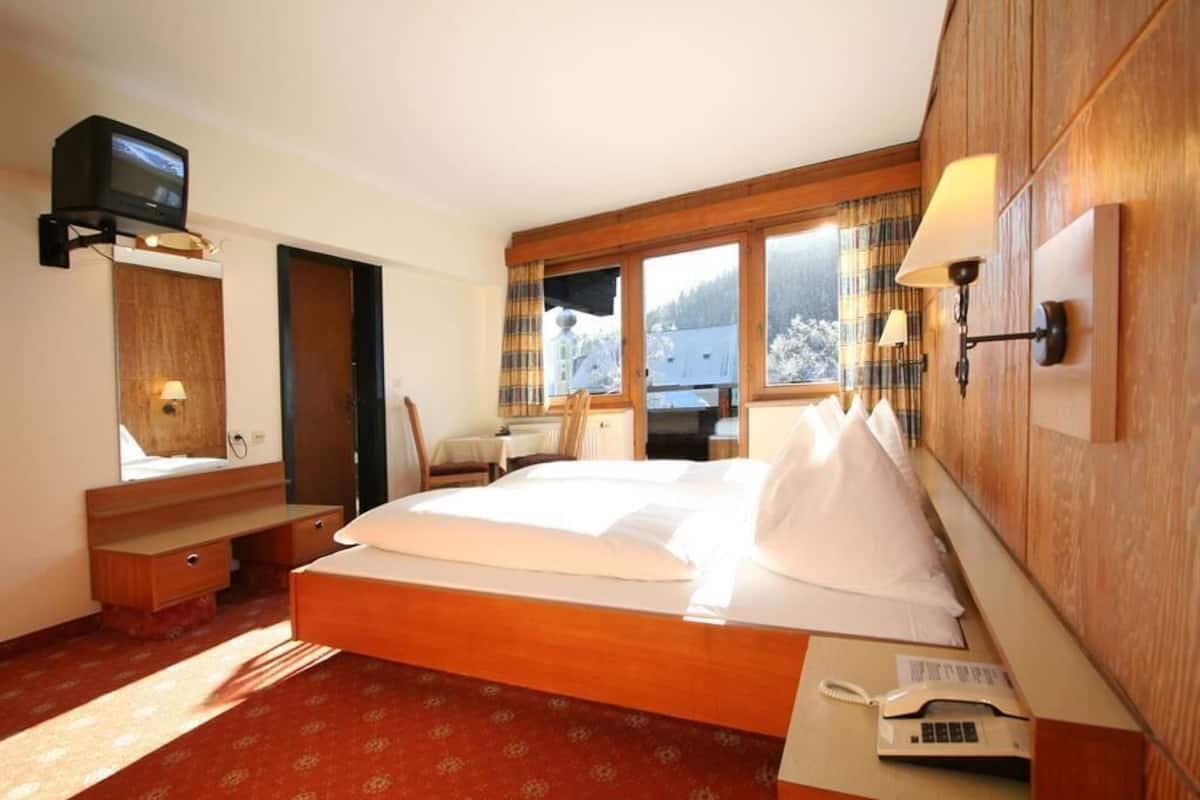 A hotel room with a wooden bed, a television mounted on the wall, a desk with a telephone, and a window with a view of a snowy landscape.