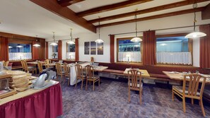 Un espace repas avec des tables et des chaises en bois, une moquette à motifs et de grandes fenêtres avec des rideaux.