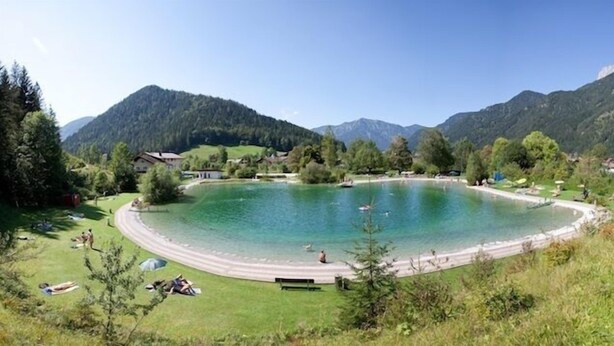 Un lago circular rodeado de casas y árboles, con montañas al fondo.