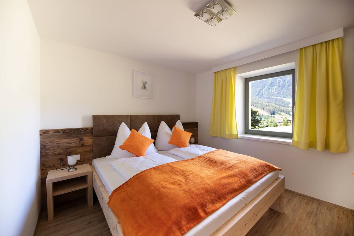 A bedroom with a wooden headboard, a large bed with white and orange bedding, a bedside table with a lamp, and a window with yellow curtains.