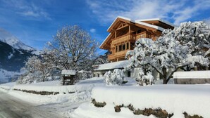 A wooden chalet with a balcony, surrounded by snow-covered trees and a clear sky.