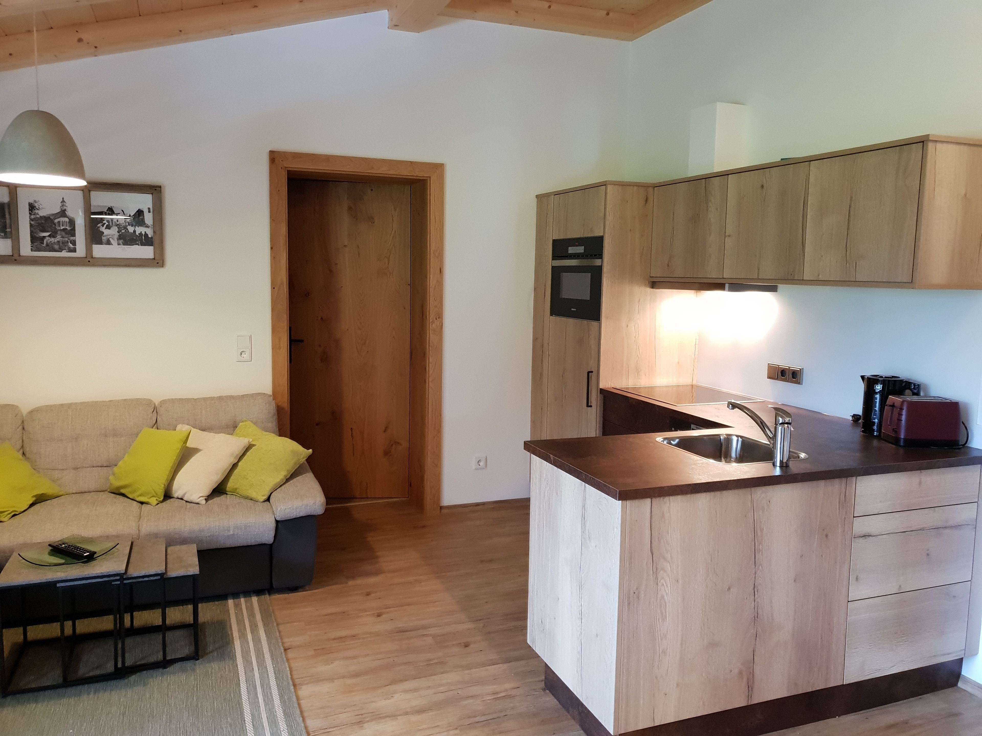 A compact living space with a kitchenette, a wooden door, a sofa with cushions, and a small table.