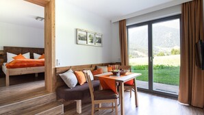 A modern room with a wooden floor, a sofa with orange cushions, a dining table with chairs, and a large window with a view of greenery and mountains.