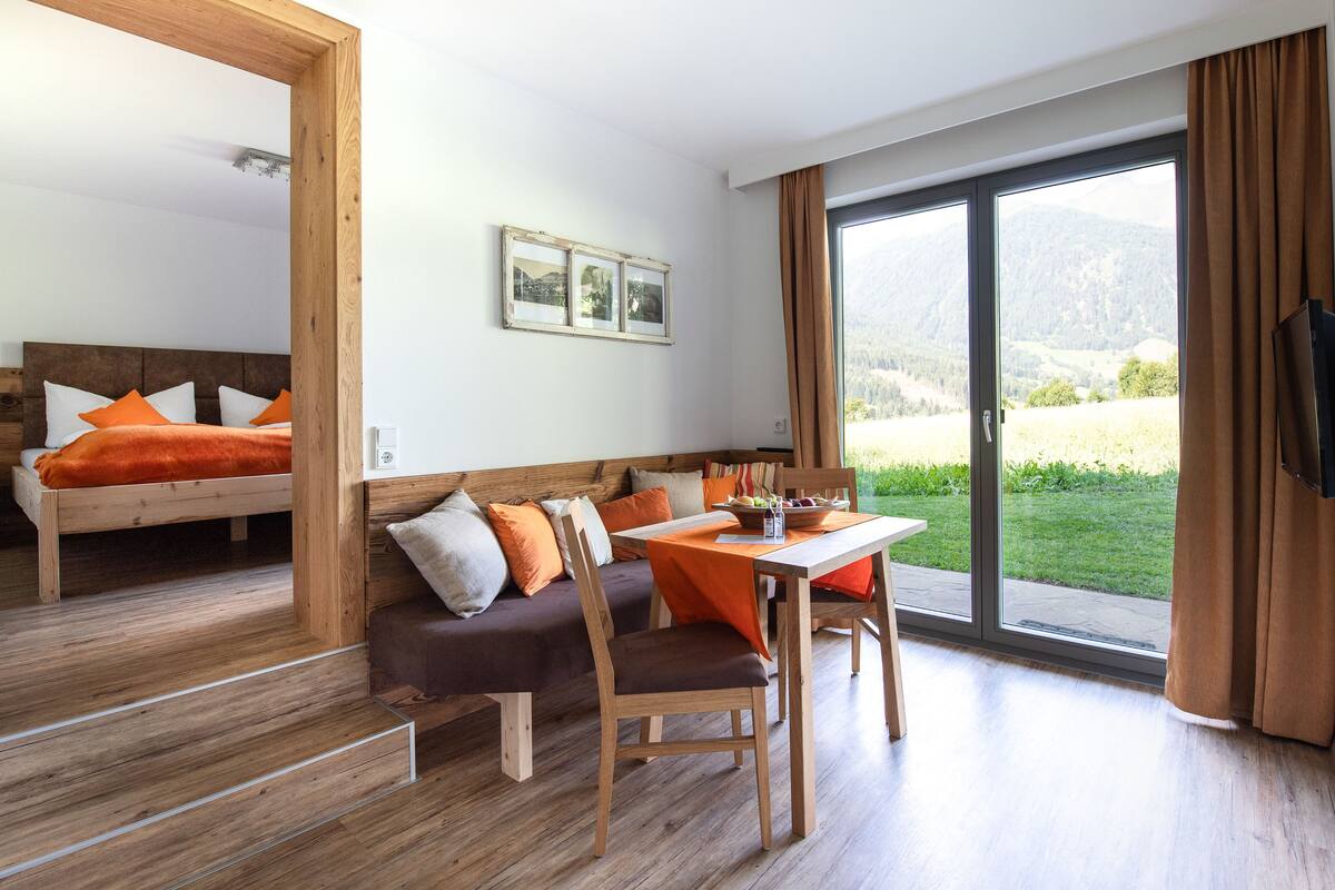 A modern room with a wooden floor, a sofa with orange cushions, a dining table with chairs, and a large window with a view of greenery and mountains.