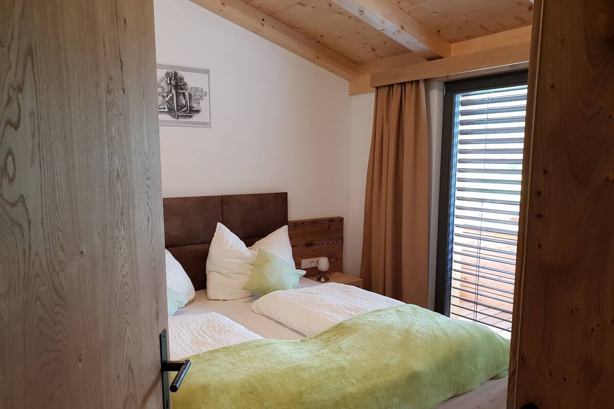 A bedroom with a wooden headboard, a bed with white and green bedding, a nightstand with a lamp, and a window with blinds.