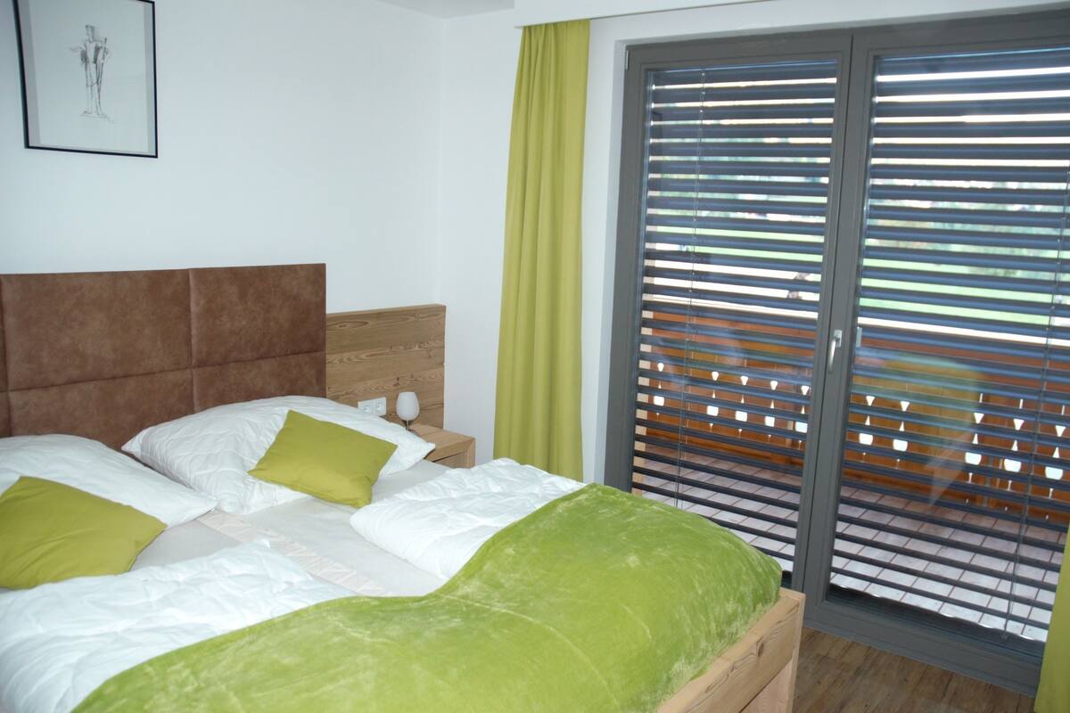 A bedroom with a bed, green pillows, a wooden headboard, a nightstand with a lamp, and a window with blinds.
