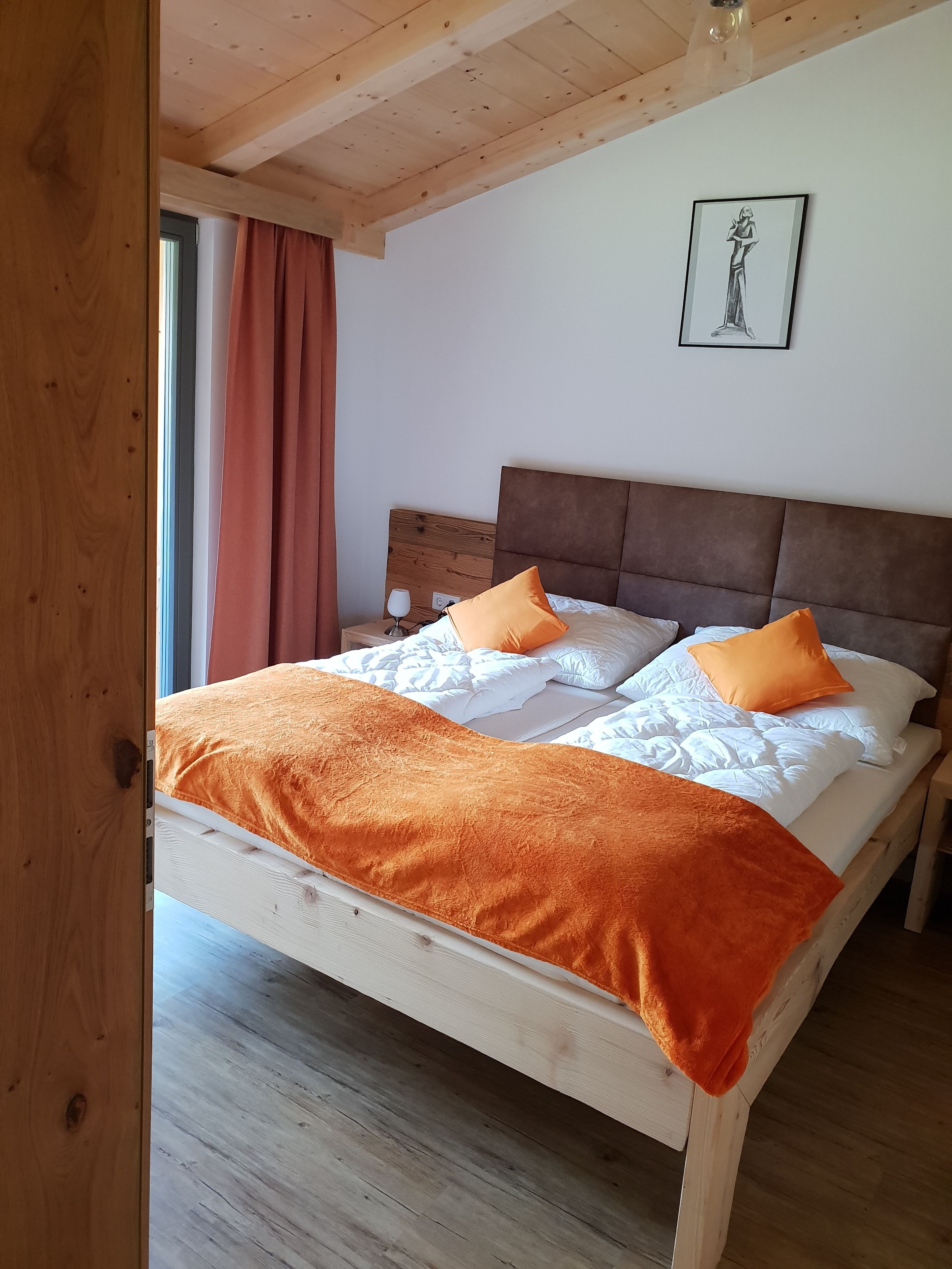 Una cama de madera con manta y almohadas anaranjadas, un cabecero y un cuadro en la pared.