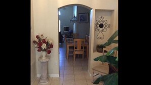 Un couloir mène à un espace repas comprenant une table et des chaises, une plante en pot et un élément décoratif fixé au mur.