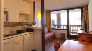 A compact kitchen with white cabinets, a sink, and a small refrigerator. There is a dining table with chairs and a bench with a cushion. A lamp is on a shelf, and a window with curtains is visible.