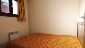 A small, single-bed room with a patterned bedspread, a window with curtains, a radiator, and a wall-mounted light fixture.