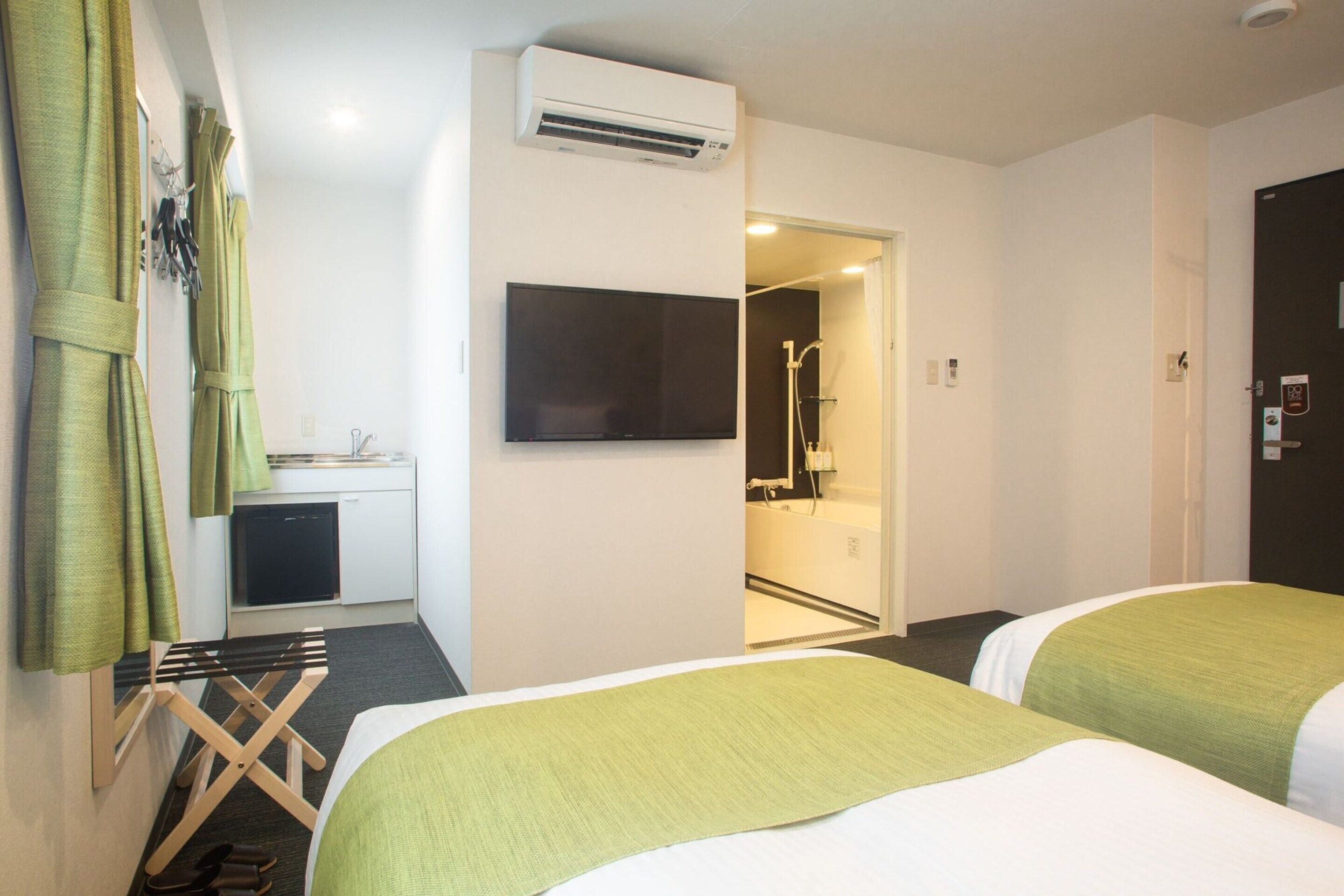A hotel room with two beds, a TV, an air conditioner, a small kitchenette, and a shower area.