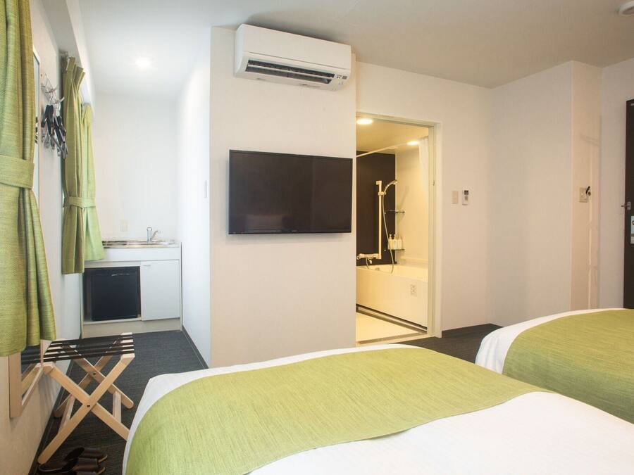 A hotel room with two beds, a TV, an air conditioner, a small kitchenette, and a shower area.