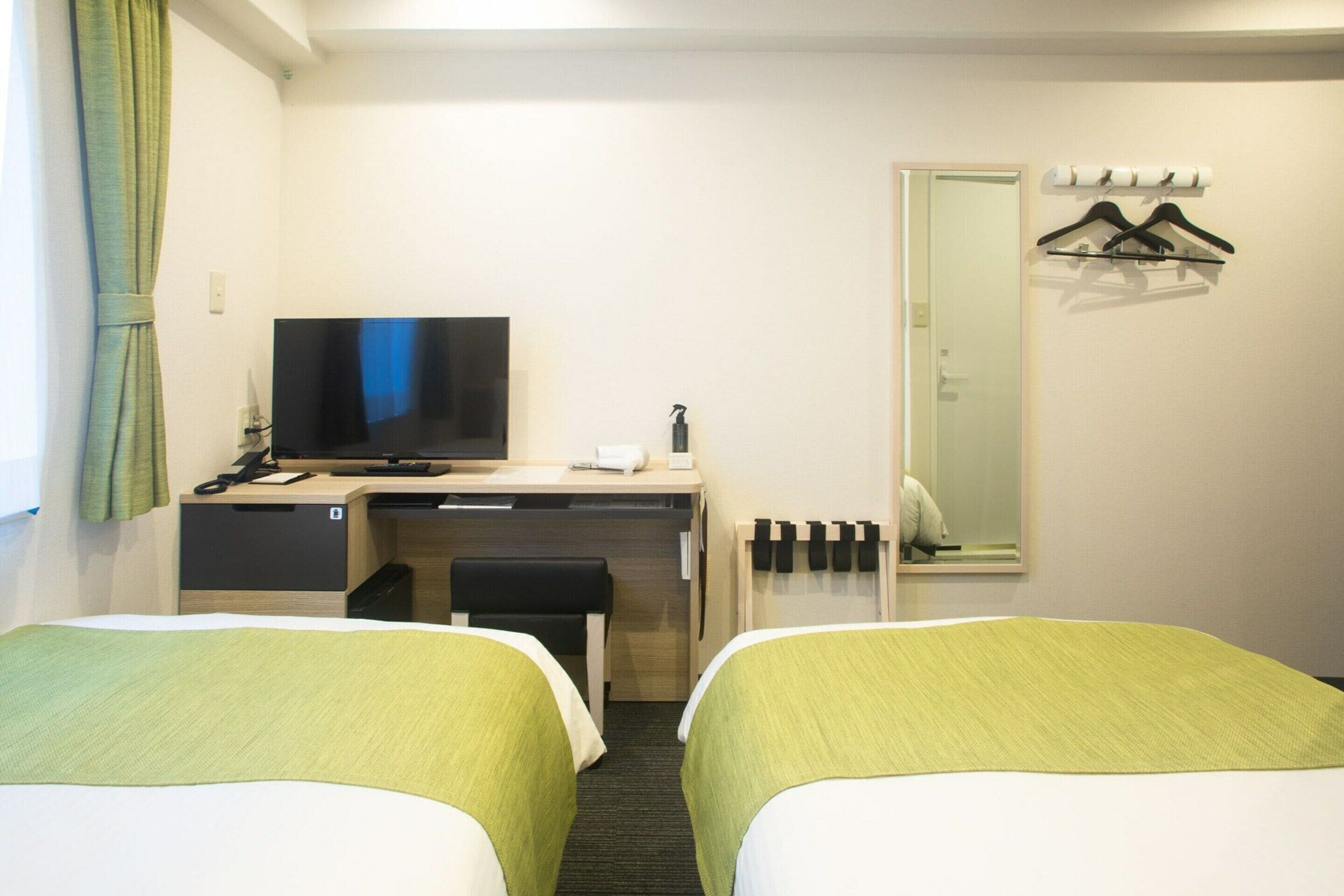 A hotel room with two beds, a desk with a television, a mirror, and a coat rack.