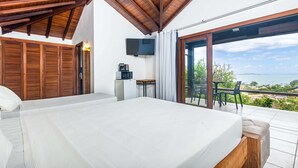 Une chambre avec un grand lit, des poutres apparentes en bois au plafond, une télévision, une petite table avec une machine à café et un balcon avec vue sur la mer.