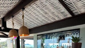 Een bar met een stenen toonbank, houten stoelen, hangende rieten lampen en uitzicht op zee.