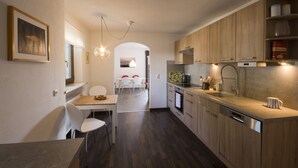 Una cocina moderna con armarios de madera, horno empotrado y lavavajillas. Hay un comedor con una mesa y sillas blancas.