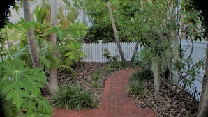 Un vialetto in mattoni che si apre su un giardino con piante tropicali e un recinto bianco in legno in secondo piano.