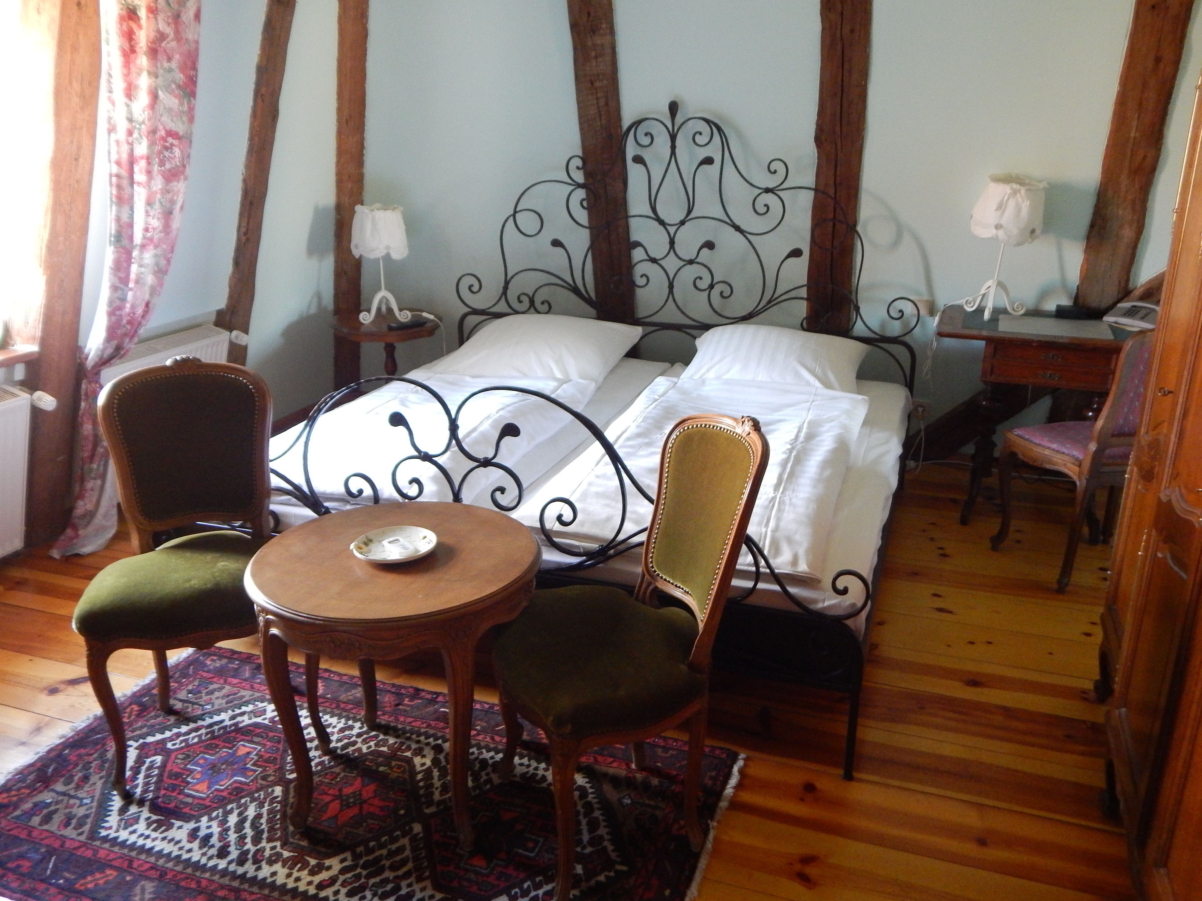 A bedroom with a bed, two chairs, a small table, a nightstand, and a desk with a lamp.