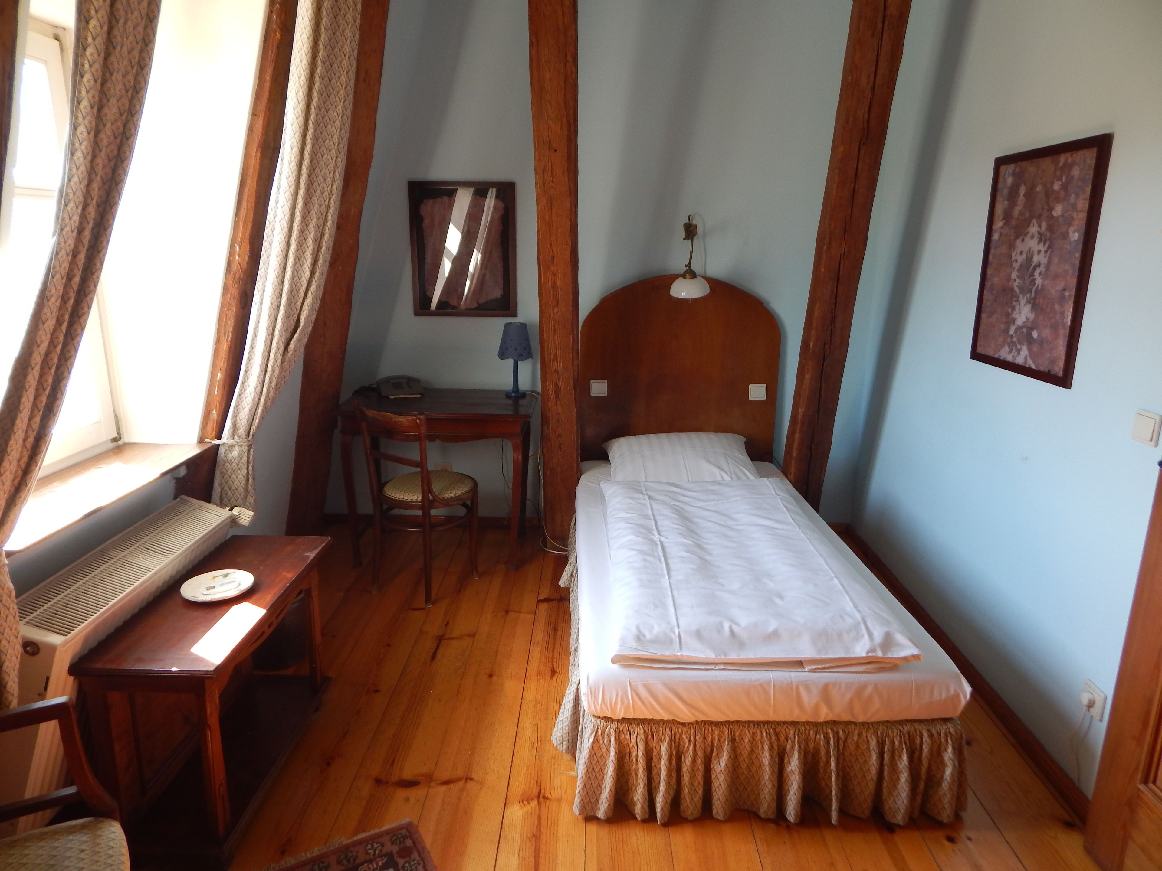 A small, cozy room with a wooden desk, a chair, a bed, and a window with curtains.
