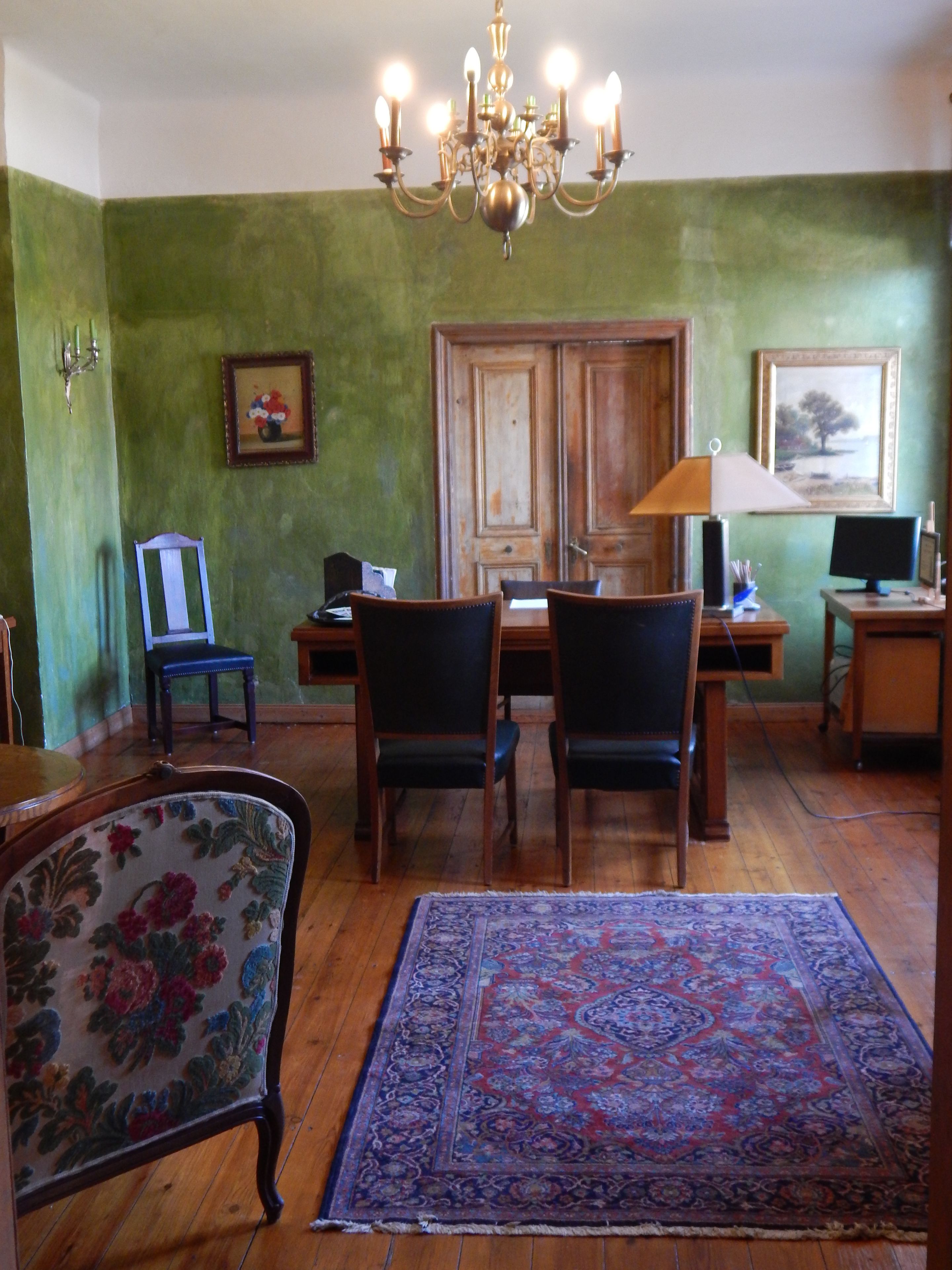 Una stanza con un tavolo da pranzo in legno, quattro sedie, un lampadario, una porta in legno, un quadro incorniciato, una lampada e un computer su una scrivania.