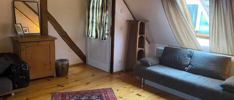 A cozy loft bedroom with a sloped ceiling, wooden beams, a gray sofa, a small cabinet, and a patterned rug.