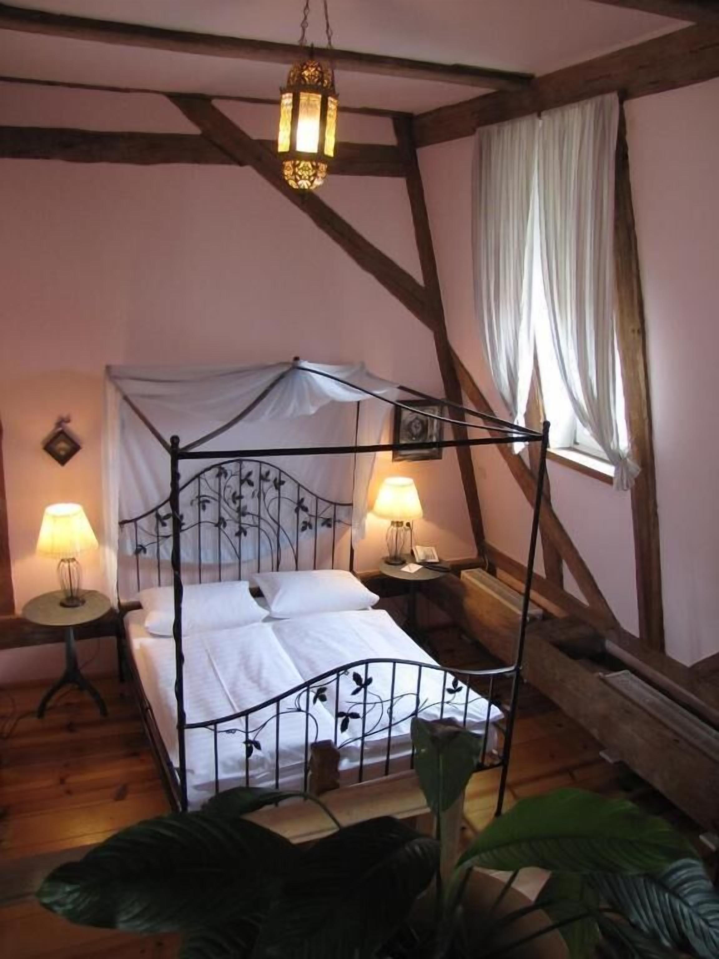 A four-poster bed with a canopy, two bedside lamps, and a hanging lantern in a room with wooden beams and a window with sheer curtains.