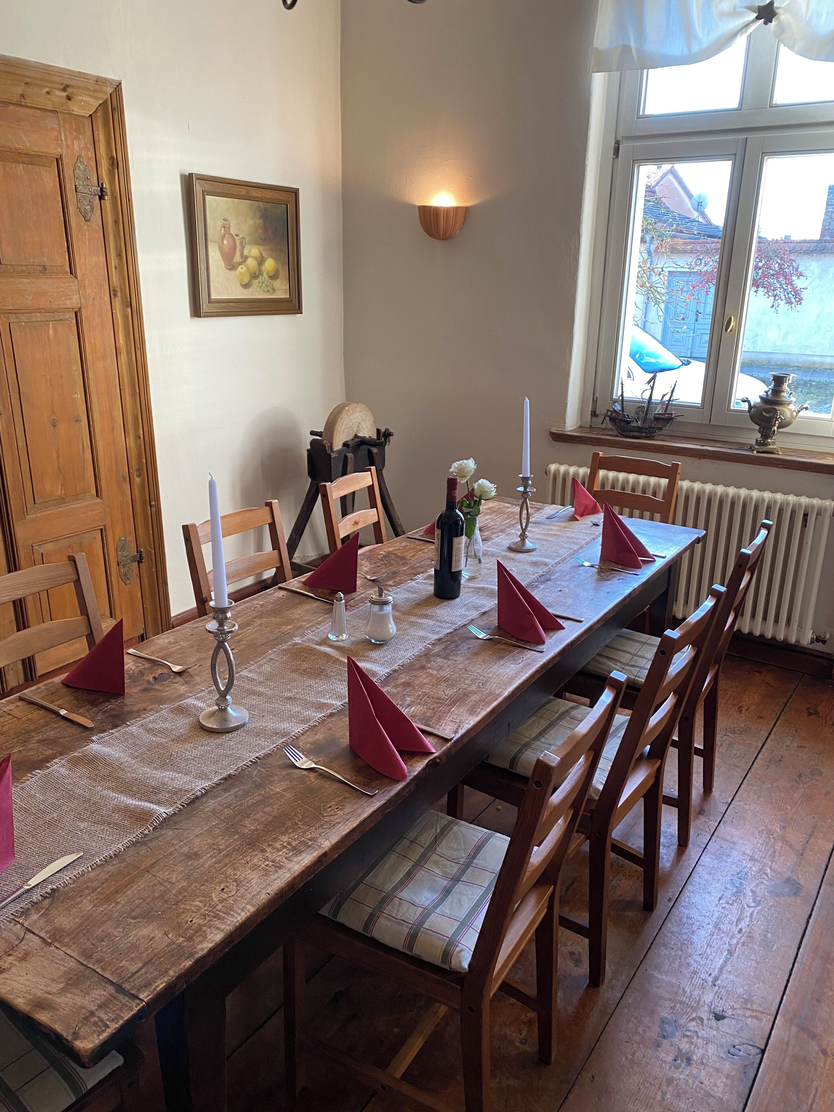 Une longue table à manger en bois, dressée avec des serviettes rouges, des bougies et une bouteille de vin, entourée de chaises en bois.