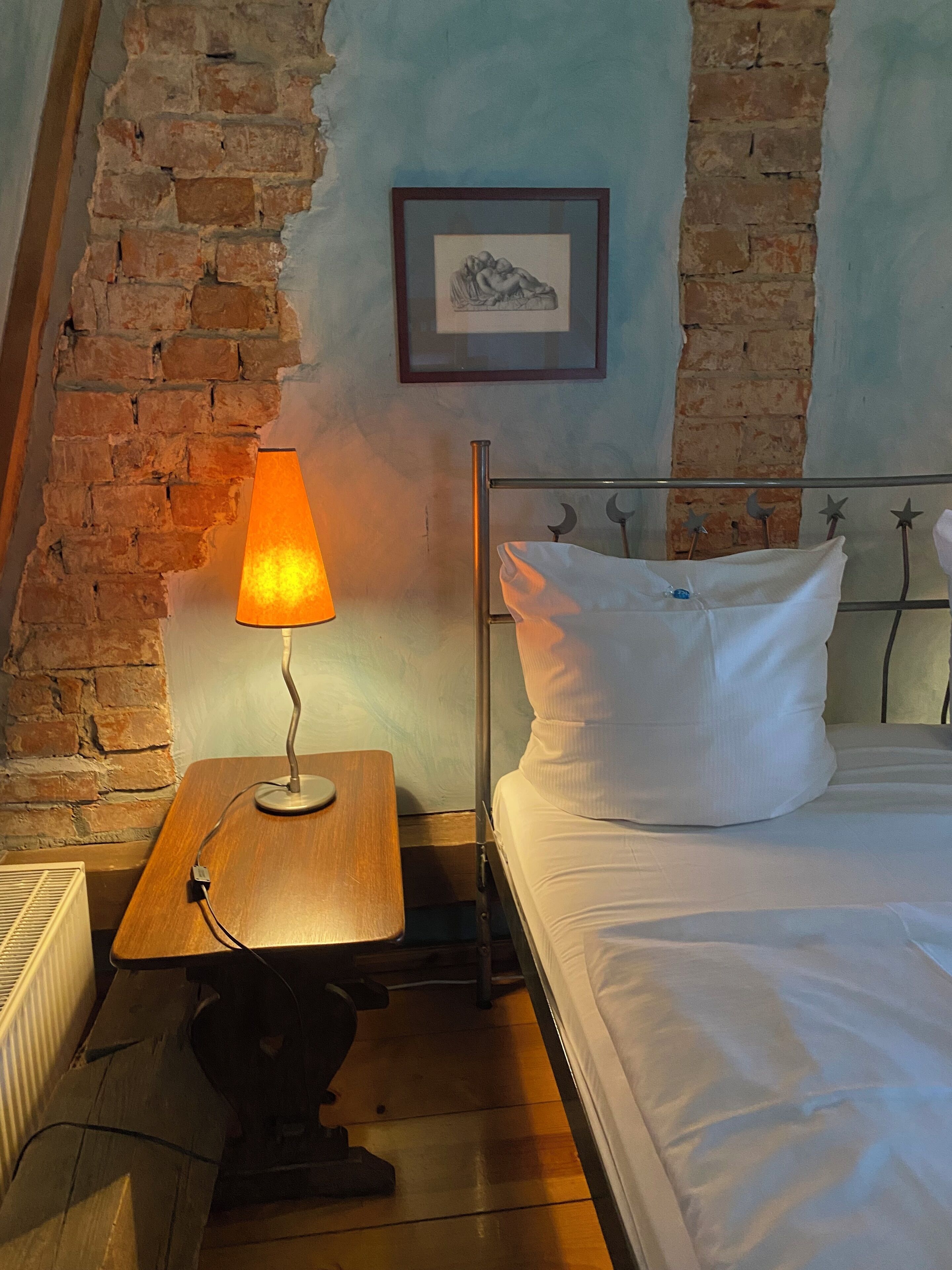 A bedroom with a brick wall, a framed picture, a bedside table with a lamp, a bed with white bedding, and a radiator.