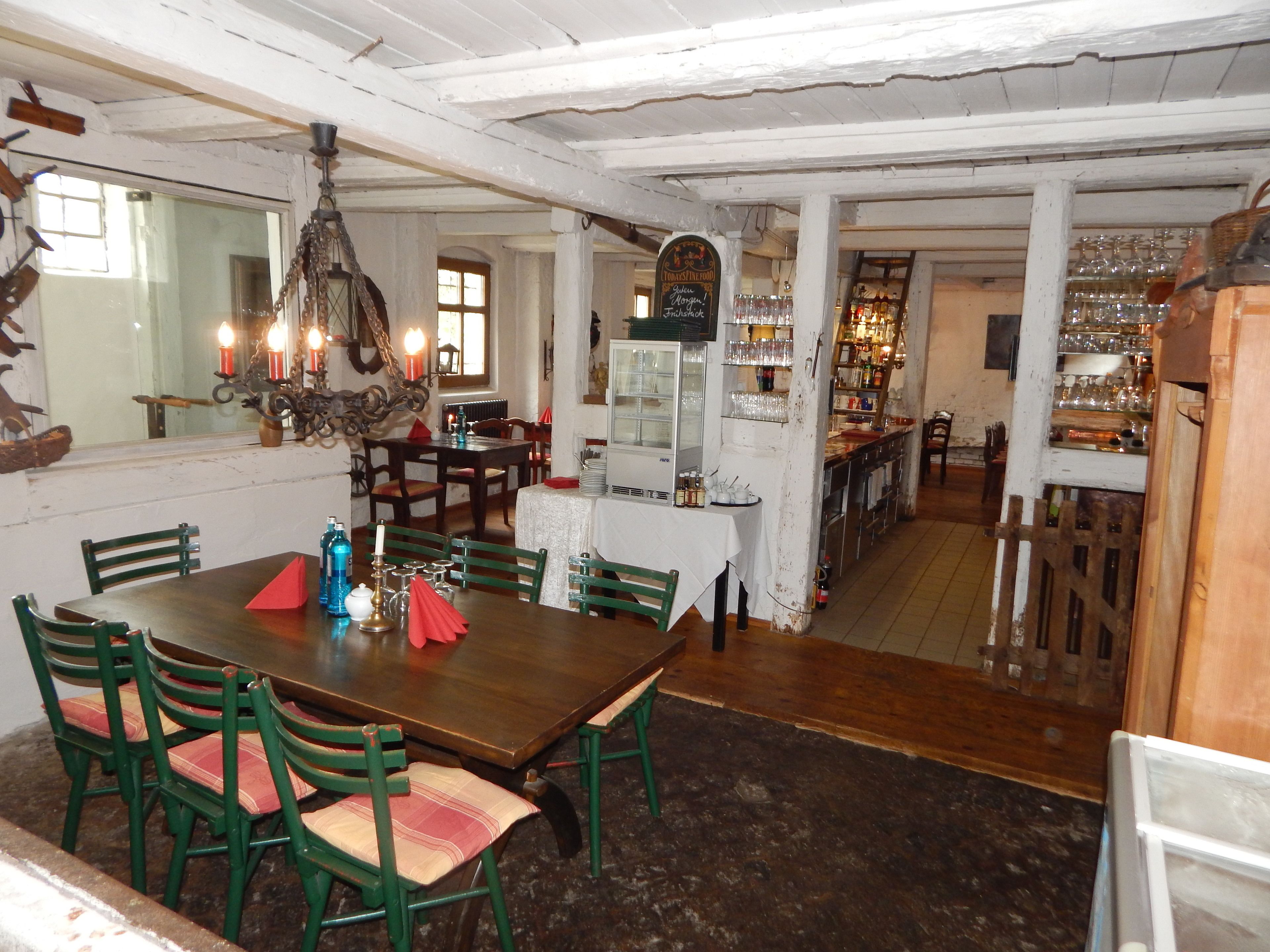 Ein Essbereich mit einem Holztisch und grünen Stühlen, einer Chandelierleuchtre und einer Barbereich mit Regalen voller Gläser und Flaschen.