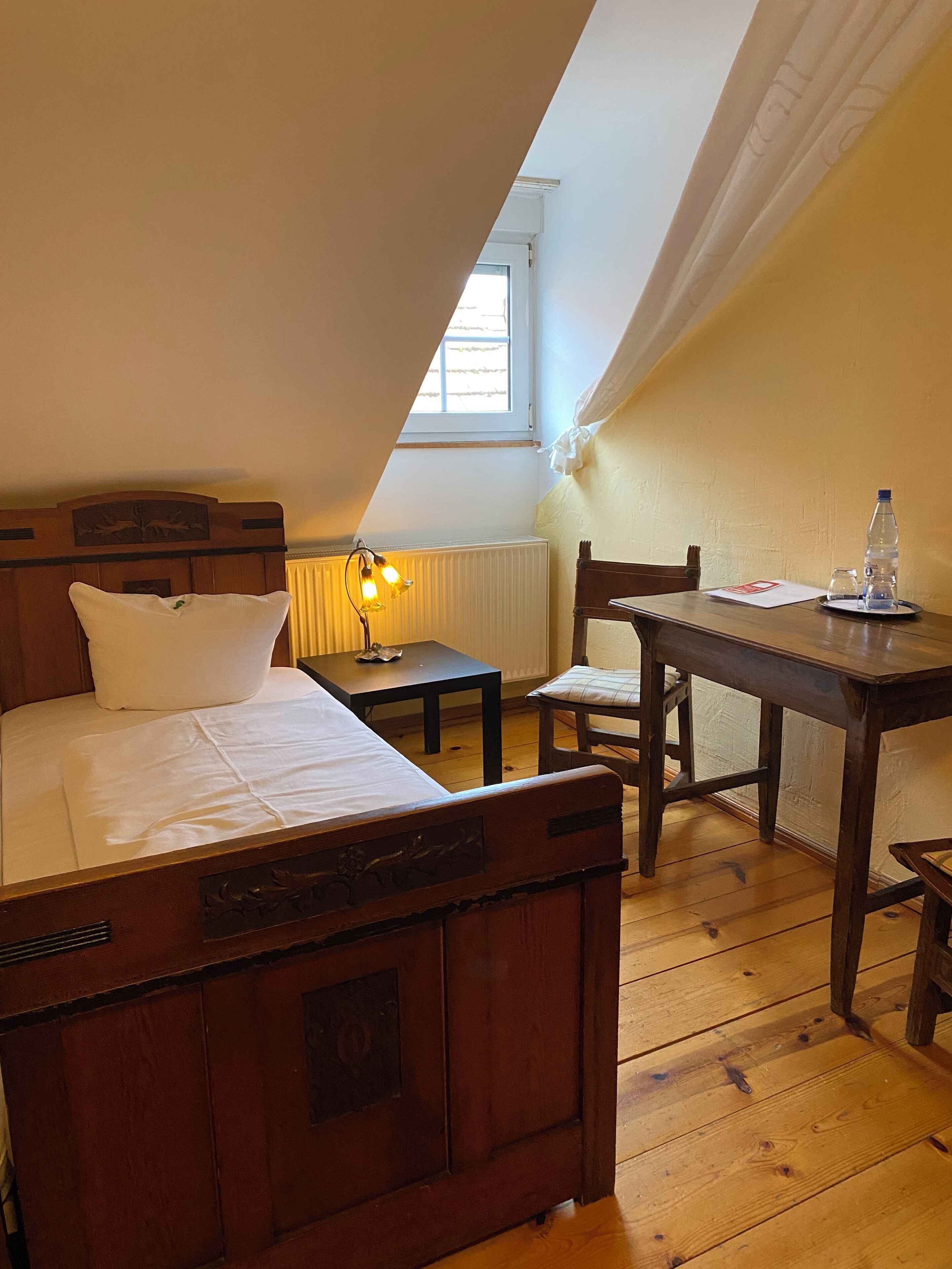 A bedroom with a wooden bed, a small table with a lamp, a chair, and a window with curtains.