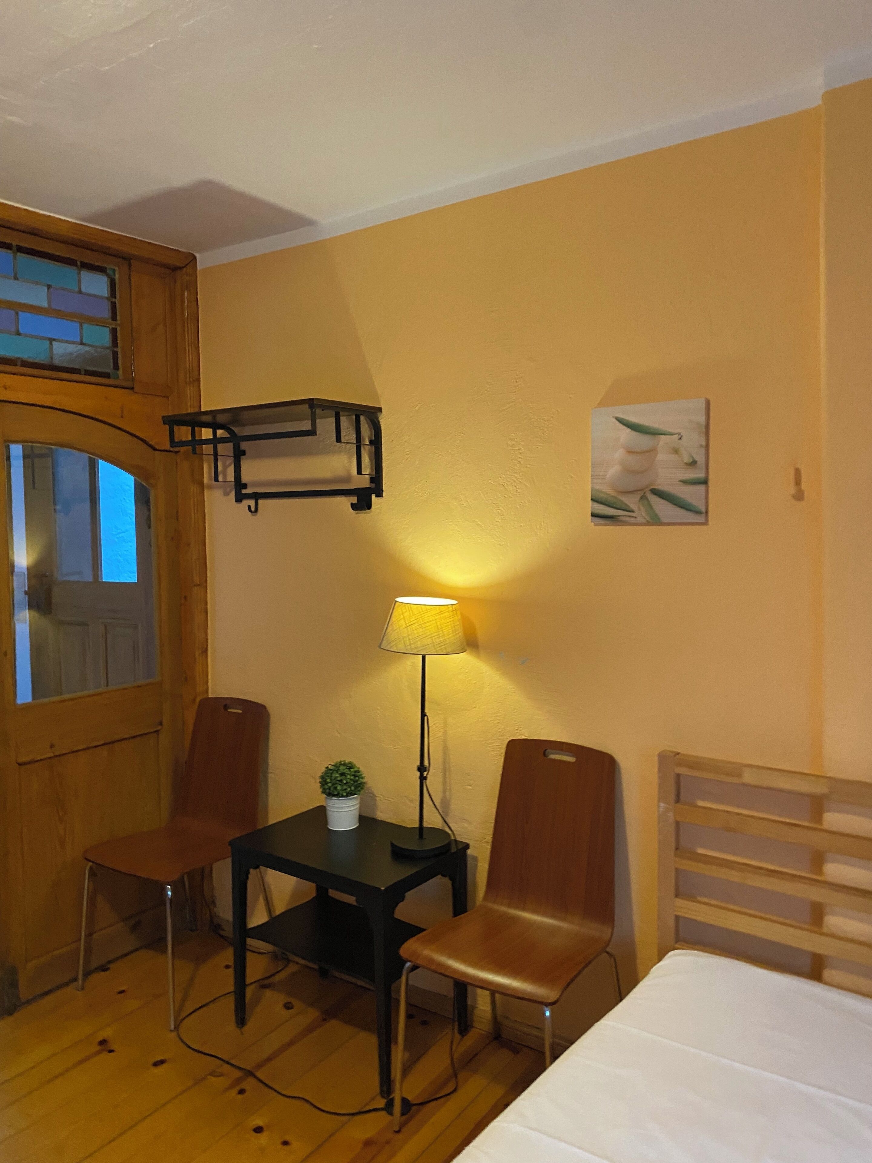 A room with a bed, two chairs, a small table with a lamp, a potted plant, and a picture on the wall.