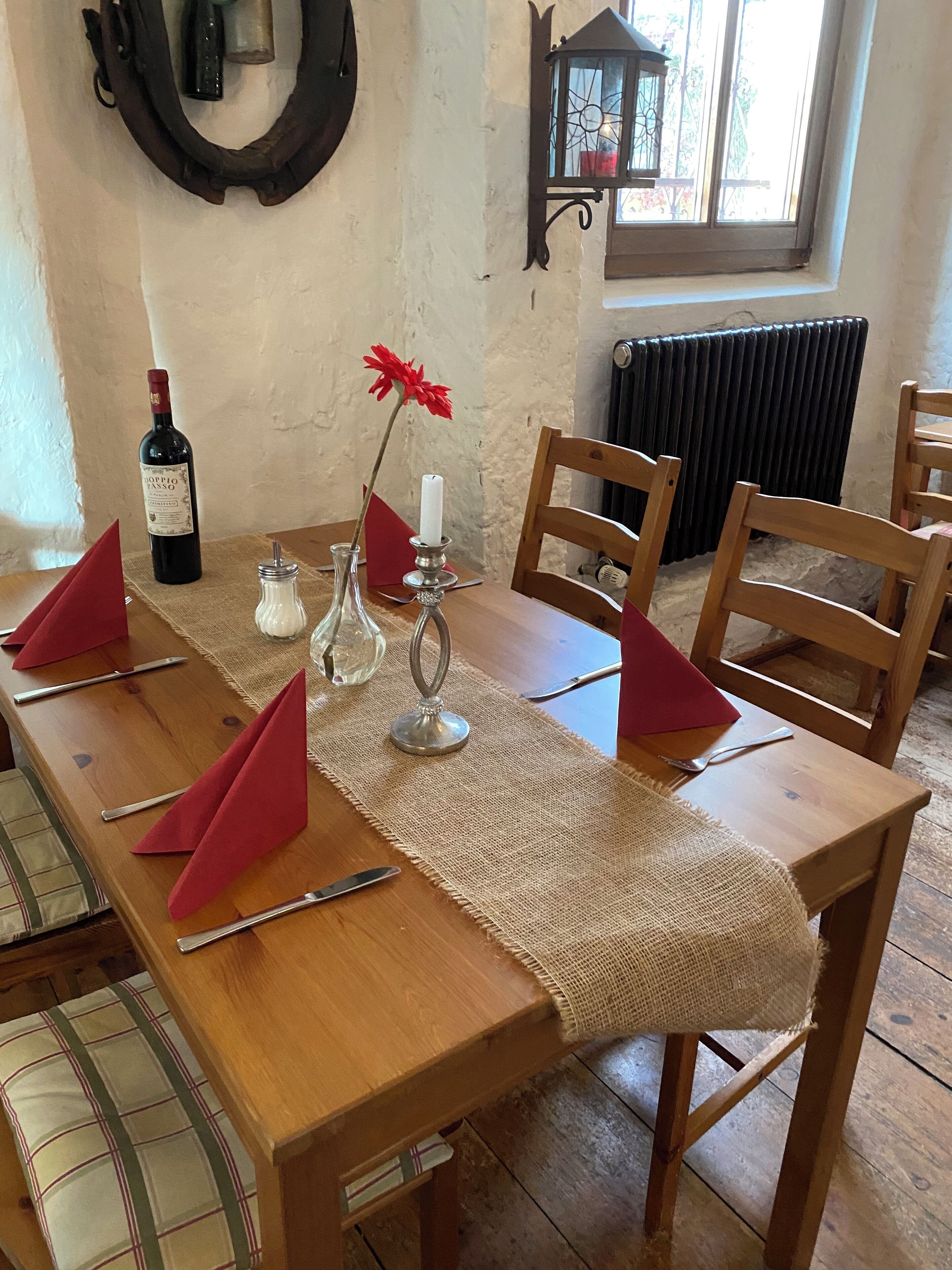 A wooden dining table set with red napkins, a bottle of wine, a candle, and a vase with a red flower.